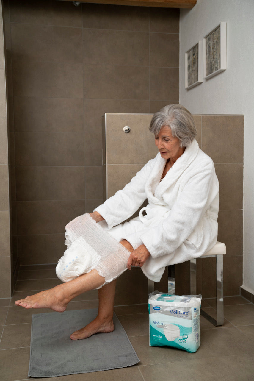 Woman in a white robe sitting on a stool in a bathroom with a package of卫生纸 on the floor.