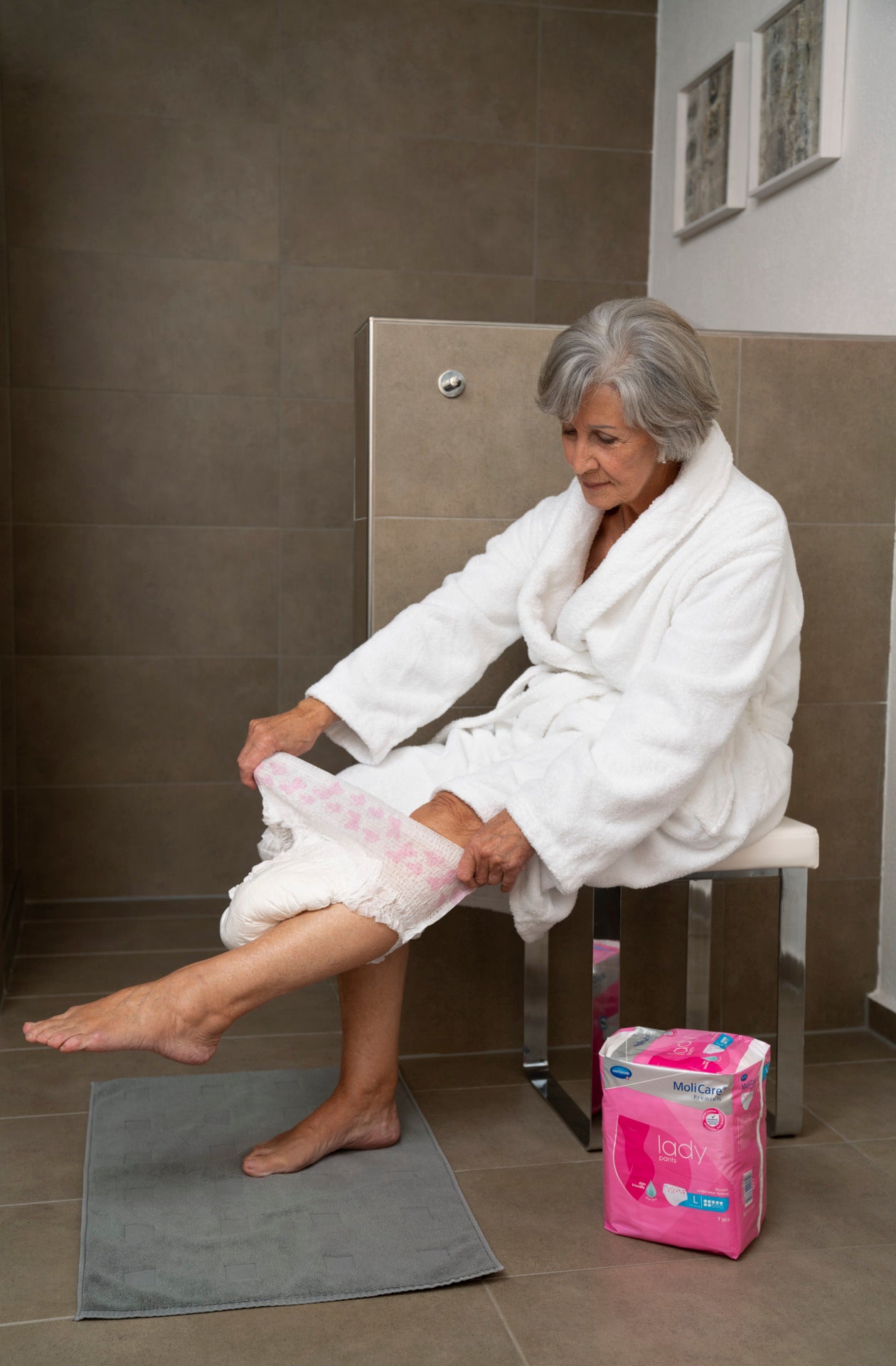 Woman in a white robe sitting on a toilet with a pink package of卫生巾 next to her.