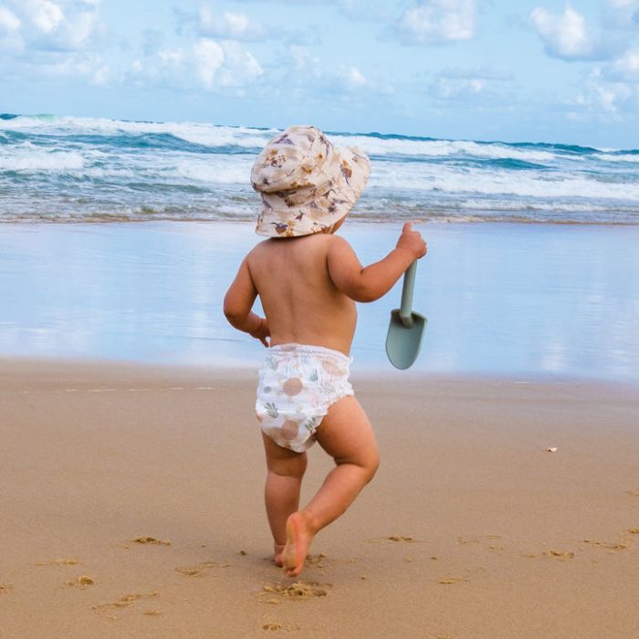 Child running on a beach with a shovel, wearing a hat and diaper.
