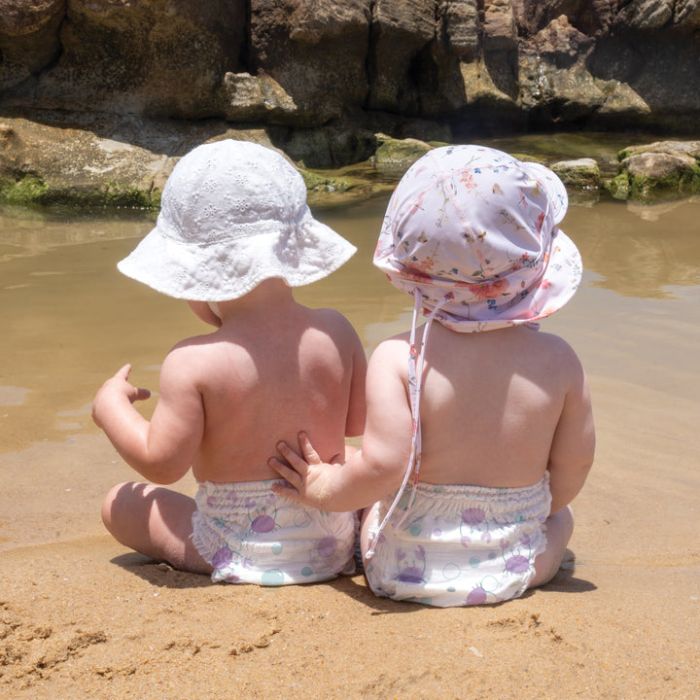 Two children sitting on a sandy beach wearing sunhats.