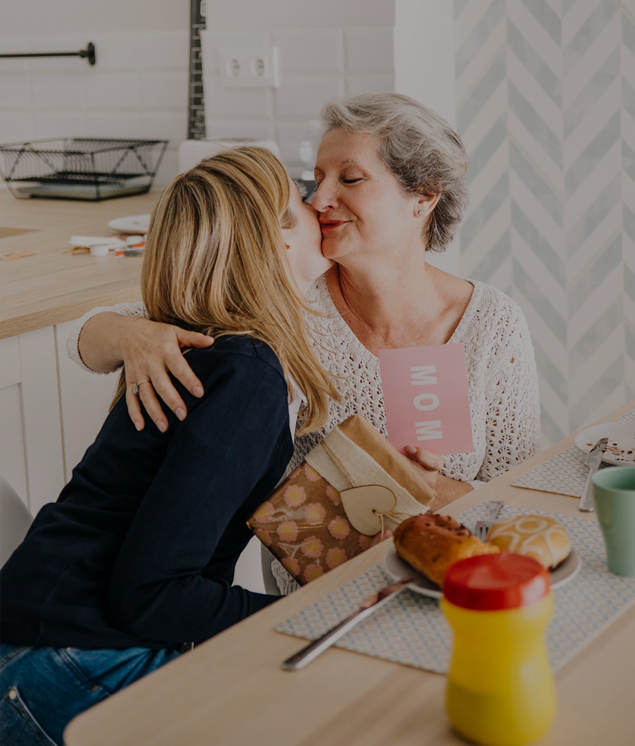 Woman hugging an older woman at a kitchen table with a 'Mom' card.