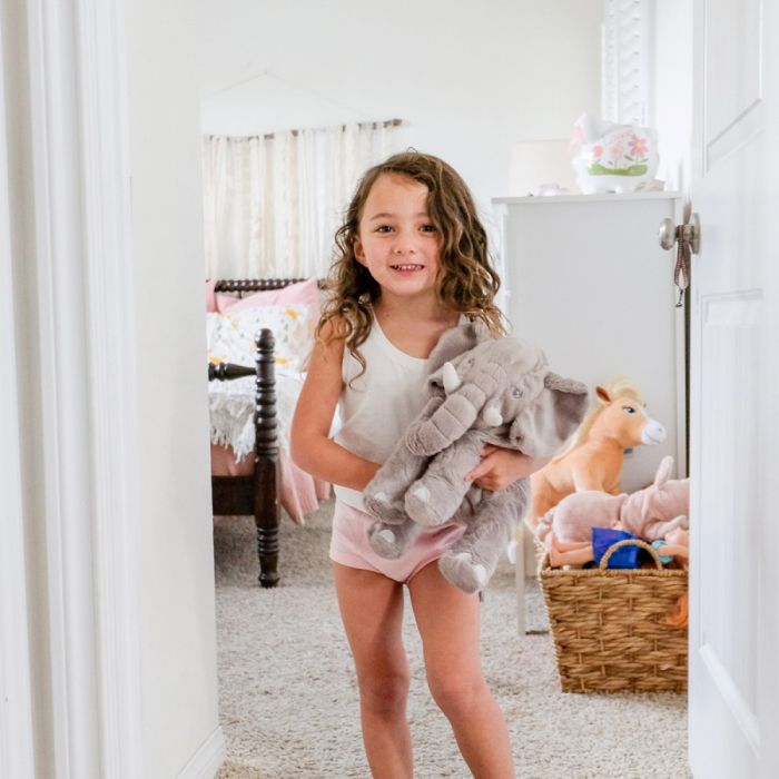 Young girl holding a plush toy in a bedroom with a basket of toys on the floor.